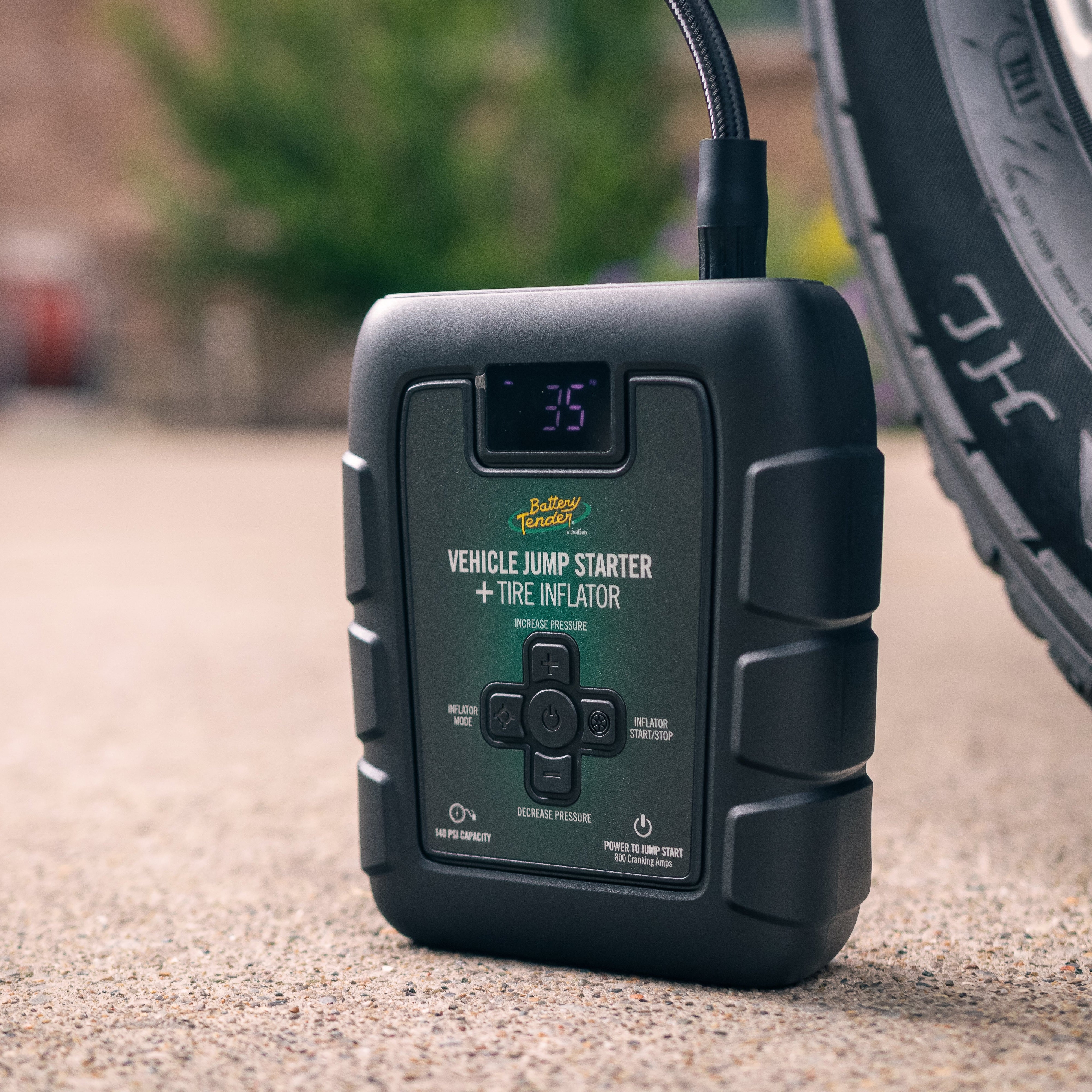 Portable vehicle jump starter and tire inflator on a concrete surface with a blurred tire in the background.